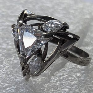 May include: A silver-toned ring featuring a central triangular-cut clear gemstone flanked by two smaller marquise-cut clear stones. The metal band has an open, intricate, and textured design that frames the three stones in a triangular arrangement.