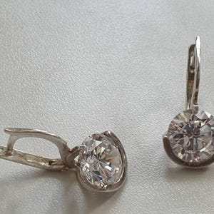 Puede incluir: Un par de pendientes de plata con cierre de palanca. Cada pendiente presenta una gran gema redonda y transparente engastada en un bisel de plata. Los pendientes se muestran sobre una superficie gris claro.