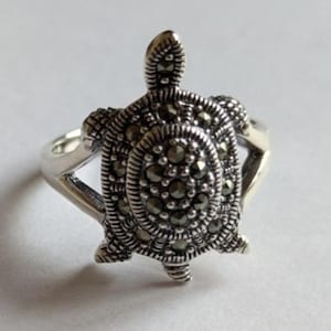 Puede incluir: Un anillo plateado con un diseño de tortuga. El caparazón de la tortuga está detallado con pequeñas piedras oscuras, y la banda del anillo es lisa y pulida. También se ven la cabeza y las aletas de la tortuga.