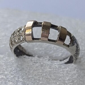 Peut inclure: Une bague argentée composée d'un anneau avec une finition texturée et tachetée de sombre sur un côté, et trois barres verticales dorées, lisses et surélevées sur le dessus, à côté d'une section incrustée de petites pierres rondes et claires.