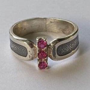 Könnte beinhalten: Ein silberner Ring mit drei rosa Edelsteinen, die vertikal angeordnet sind. Der Ring hat ein strukturiertes, dunkelgraues Banddesign auf beiden Seiten. Die Edelsteine haben eine leuchtend rosa Farbe.