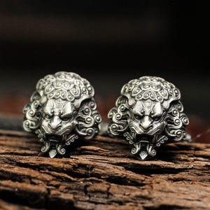 May include: Two silver-colored rings, each featuring a detailed lion's head design. The rings are placed on a piece of dark, textured wood, with a blurred background. The lion heads have intricate details, including a mane and facial features.