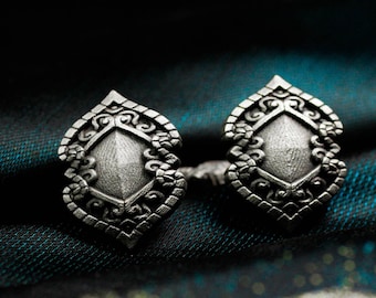 May include: Two silver-toned stud earrings with an ornate, symmetrical design. Each earring features a central diamond shape surrounded by detailed scrollwork and a decorative border. The earrings are displayed on a dark, patterned fabric.