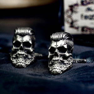 Op de afbeelding: Twee zilveren schedel manchetknopen met een baard en een kruis op het voorhoofd. De manchetknopen liggen op een donkerblauwe stoffen achtergrond.