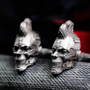 Op de afbeelding: Een paar zilverkleurige manchetknopen, elk in de vorm van een schedel met een hanenkam. De manchetknopen zijn gedetailleerd met gezichtskenmerken en staan tegen een donkere achtergrond.