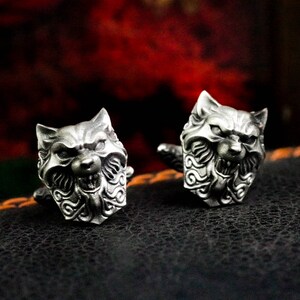 May include: A pair of silver cufflinks featuring a detailed wolf head design. The wolf's mouth is open, showing sharp teeth, and its fur is intricately sculpted.