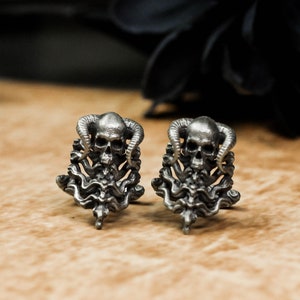 May include: A pair of silver skull cufflinks with intricate details. Each skull is adorned with horns and surrounded by a swirling design.