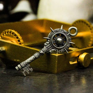 May include: An ornate, silver-toned key pendant with a sunburst design. The key features detailed patterns on the handle and head. It is placed on a gold-coloured box, with a skull visible in the background.