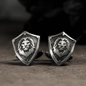 Puede incluir: Un par de gemelos plateados. Cada gemelo presenta un diseño de escudo con la cabeza de un león detallada en el centro. Los gemelos se muestran sobre un fondo oscuro, resaltando los intrincados detalles de la melena y los rasgos faciales del león.