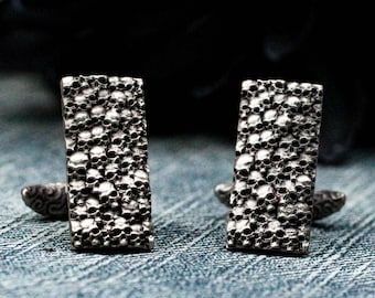 Puede incluir: Un par de gemelos de plata con un patrón de calavera. Los gemelos son de forma rectangular y tienen una superficie texturizada.