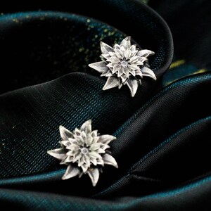 May include: A pair of silver flower stud earrings. The earrings have a detailed petal design and are set against a dark, textured background.