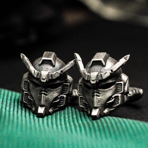May include: A pair of silver robot head cufflinks. The cufflinks have a detailed, angular design and a metallic finish. They are displayed on green fabric, set against a dark background.