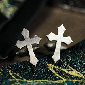 May include: A pair of silver-colored cross-shaped earrings. The earrings have a gothic design with pointed edges. They are displayed on a textured, dark blue and gold patterned surface.
