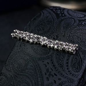 以下が含まれることがあります： 骸骨のデザインが施されたシルバーのネクタイピン。ネクタイピンは、黒のペイズリー柄のネクタイの上に置かれています。