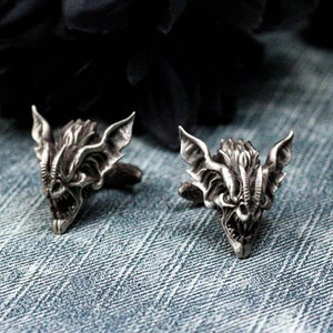 May include: A pair of silver cufflinks featuring a detailed bat design with sharp teeth and wings. The cufflinks are on a blue denim background.