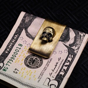 Peut inclure: Un pince-billets en laiton avec un motif de crâne maintient un billet de cinq dollars. Le clip est rectangulaire avec un crâne détaillé sur le devant. Le billet est partiellement visible, avec le texte "FEDERAL RESERVE SYSTEM".