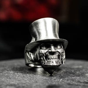 May include: A silver-colored ring featuring a detailed, gothic design. The ring depicts a skeletal face wearing a top hat. The face has intricate details, including visible teeth and facial structures. The ring is set on a dark, textured surface.