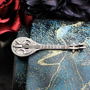 May include: A silver-colored, decorative brooch in the shape of a lute, with intricate Celtic-inspired designs. The brooch is set against a backdrop of a patterned fabric in shades of teal, gold, and black, with a dark floral arrangement in the background.