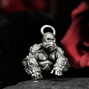 Può includere: Ciondolo a forma di gorilla color argento con superficie dettagliata e strutturata. Il ciondolo presenta un anello nella parte superiore per essere appeso, su sfondo scuro con un tocco di rosso.