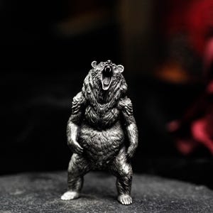 May include: A detailed silver-colored bear figurine in a standing pose with its mouth open, showcasing its teeth. The bear has a textured fur appearance and is set against a dark background, highlighting the intricate details of the sculpture.
