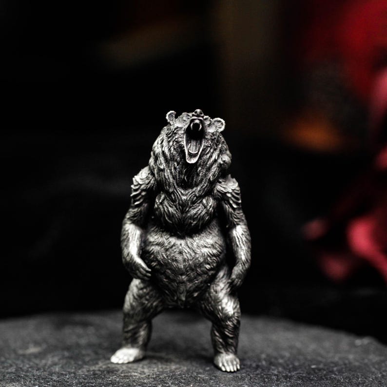 May include: A detailed silver-colored bear figurine in a standing pose with its mouth open, showcasing its teeth. The bear has a textured fur appearance and is set against a dark background, highlighting the intricate details of the sculpture.