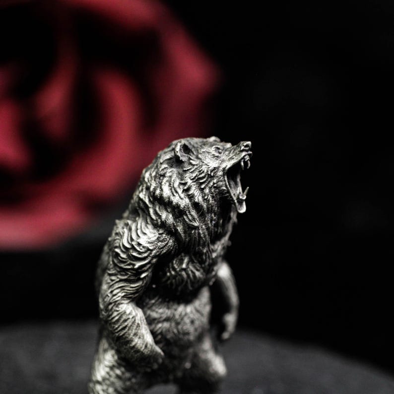 May include: A detailed silver-colored bear figurine with an open mouth, showcasing its teeth. The bear is standing upright, with textured fur and a fierce expression. The background is blurred, featuring a red rose and a dark backdrop.