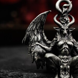 May include: A detailed, silver-toned sculpture of a seated, winged figure with a goat head, horns, and a flame-like design above the head. The figure has outstretched arms and is set against a dark background.