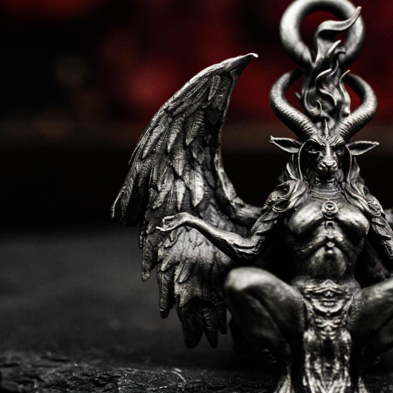May include: A detailed, silver-toned sculpture of a seated, winged figure with a goat head, horns, and a flame-like design above the head. The figure has outstretched arms and is set against a dark background.