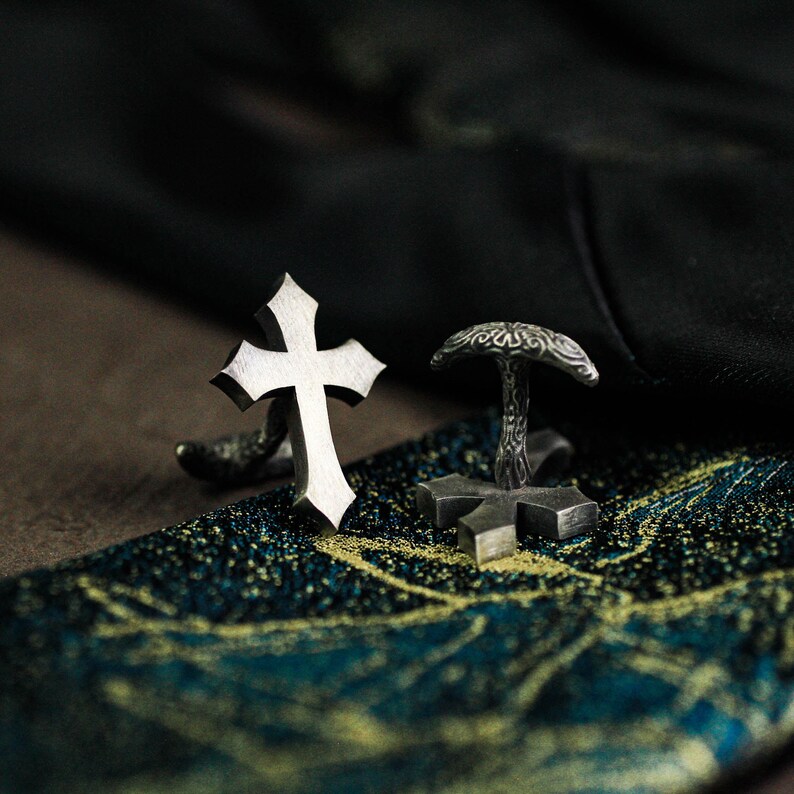 May include: Silver-toned cufflinks with a cross design. One cufflink features a detailed cross, while the other has an ornate top and a cross base. The cufflinks are displayed on a patterned surface.