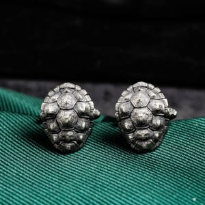 May include: A pair of silver-toned cufflinks, each shaped like a turtle shell. The cufflinks have a detailed, textured surface and a classic post closure. The background is a dark green fabric.