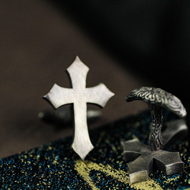 May include: Close-up of silver-toned cross-shaped cufflinks. The cufflinks have a detailed, textured design. The background is dark, highlighting the metallic sheen of the jewelry. The cufflinks are likely for formal wear.
