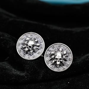 Könnte beinhalten: Zwei silberfarbene Manschettenknöpfe mit Totenkopf-Design. Jeder Manschettenknopf hat einen zentralen Totenkopf, der von kleineren Totenköpfen und römischen Ziffern am Rand umgeben ist. Die Manschettenknöpfe sind rund und haben eine detaillierte, gotische Ästhetik.