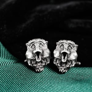May include: A pair of silver-toned cufflinks, each featuring a detailed, open-mouthed gargoyle face. The cufflinks are set against a dark background and a green fabric.