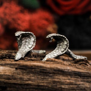 Puede incluir: Dos figuras de cobra plateadas con escamas detalladas y bocas abiertas, colocadas sobre un trozo de madera oscura. El fondo presenta elementos borrosos rojos y negros, creando un contraste dramático. Las cabezas de cobra están levantadas.