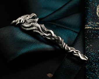 Puede incluir: Un pasador de corbata plateado con un diseño detallado de cabeza de dragón. El pasador se coloca sobre una corbata estampada en verde oscuro y negro, creando un contraste llamativo. La boca abierta y las escamas texturizadas del dragón son claramente visibles.