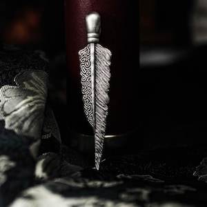 May include: A silver-colored feather pendant with intricate detailing. One side features a detailed swirl pattern, while the other side has a textured feather design. The pendant has a small, rounded top and is displayed against a dark background.