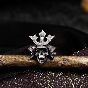 May include: A silver-colored ring featuring a skull wearing a samurai helmet. The ring is detailed with intricate carvings and sits on a wooden branch against a dark background. The skull has an open mouth and the helmet has a decorative crest.