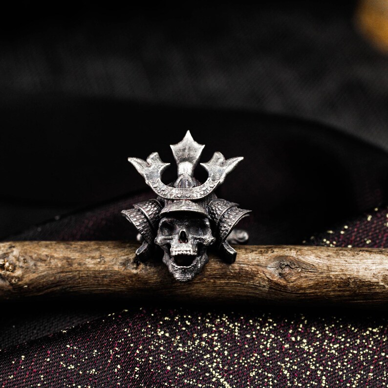 May include: A silver-colored ring featuring a skull wearing a samurai helmet. The ring is detailed with intricate carvings and sits on a wooden branch against a dark background. The skull has an open mouth and the helmet has a decorative crest.