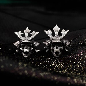 May include: A pair of silver-colored cufflinks featuring skull designs with samurai helmet details. The cufflinks are set against a dark, textured background, highlighting the intricate details of the skull and helmet.