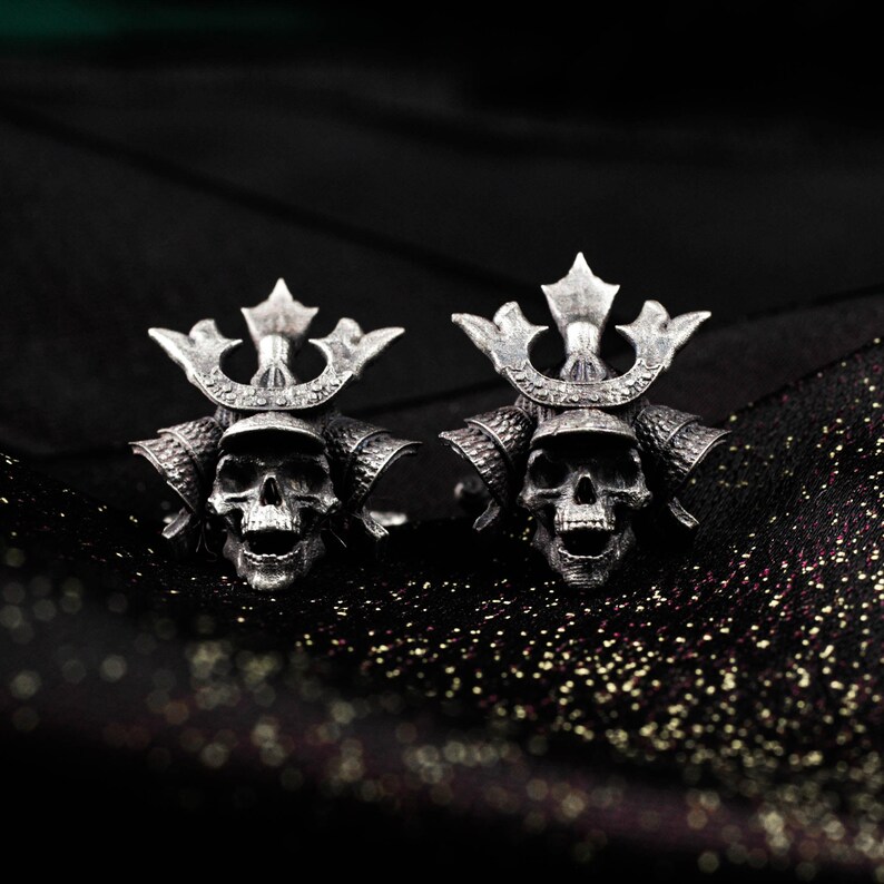 May include: A pair of silver-colored cufflinks featuring skull designs with samurai helmet details. The cufflinks are set against a dark, textured background, highlighting the intricate details of the skull and helmet.