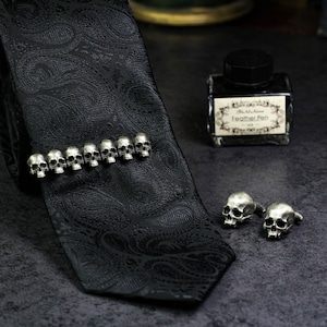 May include: A black paisley tie with a silver skull tie clip. The tie clip has nine skulls in a row. There are also two silver skull cufflinks.