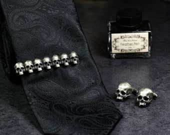 Puede incluir: Una corbata negra de paisley con un broche de corbata de calavera de plata. El broche de corbata tiene nueve calaveras en fila. También hay dos gemelos de calavera de plata.