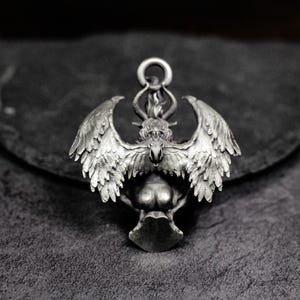 May include: A silver-toned pendant featuring a winged creature with a horned head and a flame detail. The pendant has intricate feather and wing detailing, with a loop at the top for a chain. The design is detailed and gothic in style.