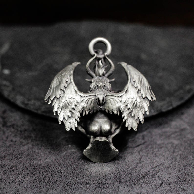 May include: A silver-toned pendant featuring a winged creature with a horned head and a flame detail. The pendant has intricate feather and wing detailing, with a loop at the top for a chain. The design is detailed and gothic in style.