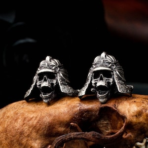 May include: Two silver skull rings with detailed samurai helmet designs. The rings are sitting on a brown, textured surface.