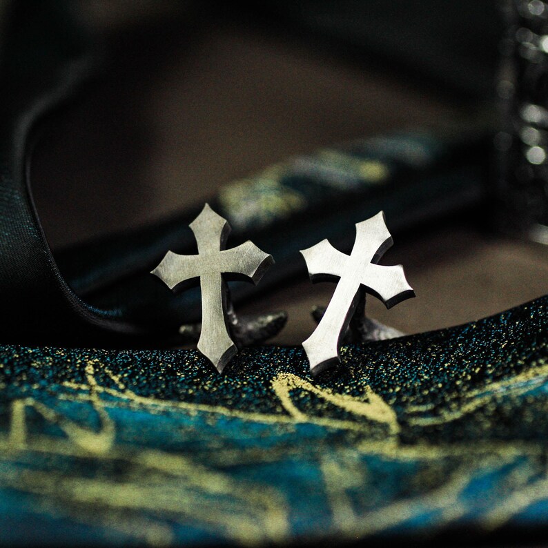 May include: A pair of silver-colored cross-shaped earrings. The earrings have a gothic design with pointed edges. The background is a dark, textured surface with teal and gold accents, creating a dramatic contrast with the earrings.