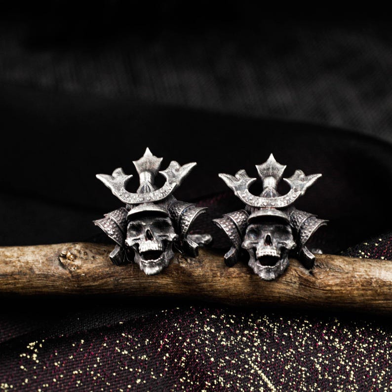 May include: Two silver-colored skull-shaped earrings, each adorned with a crown and samurai helmet details. The earrings are displayed on a wooden branch against a dark background, showcasing a gothic or biker theme.