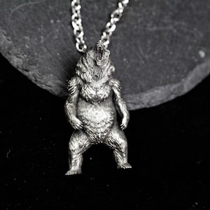 May include: A silver-colored pendant necklace featuring a detailed bear design. The bear is standing upright with its mouth open, attached to a chain. The pendant is made of a metallic material and is set against a dark background.
