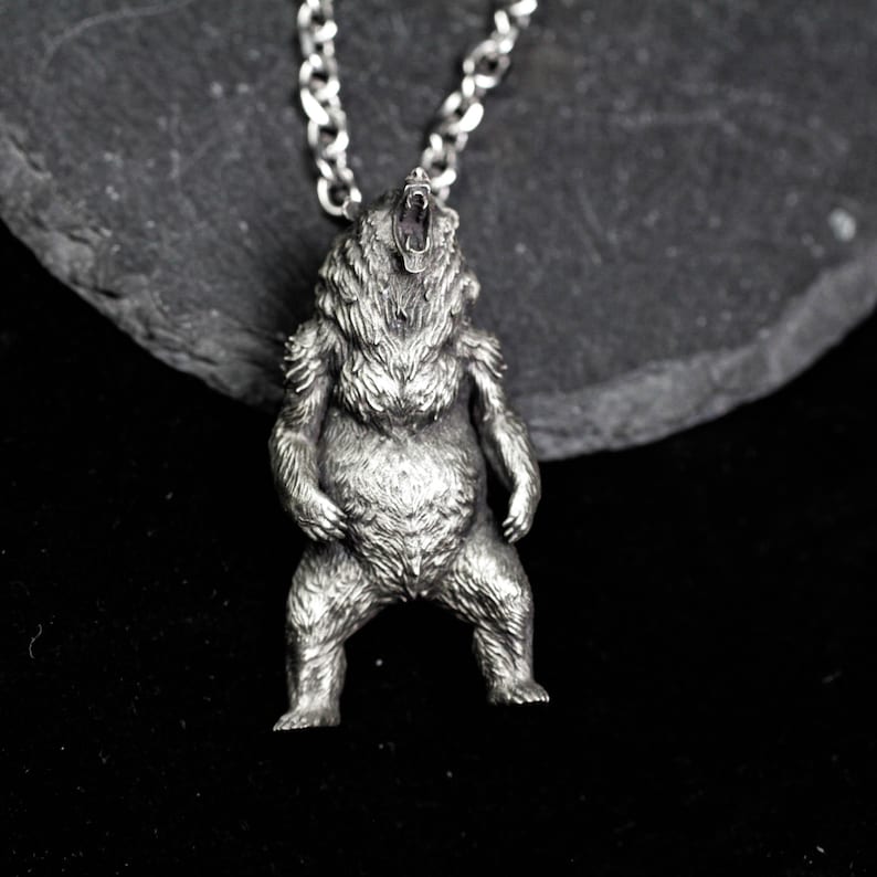 May include: A silver-colored pendant necklace featuring a detailed bear design. The bear is standing upright with its mouth open, attached to a chain. The pendant is made of a metallic material and is set against a dark background.