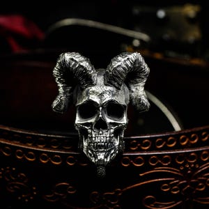 May include: A silver skull ring with large, curved horns. The skull has detailed facial features and is set against a dark background. The ring is a gothic-inspired accessory.